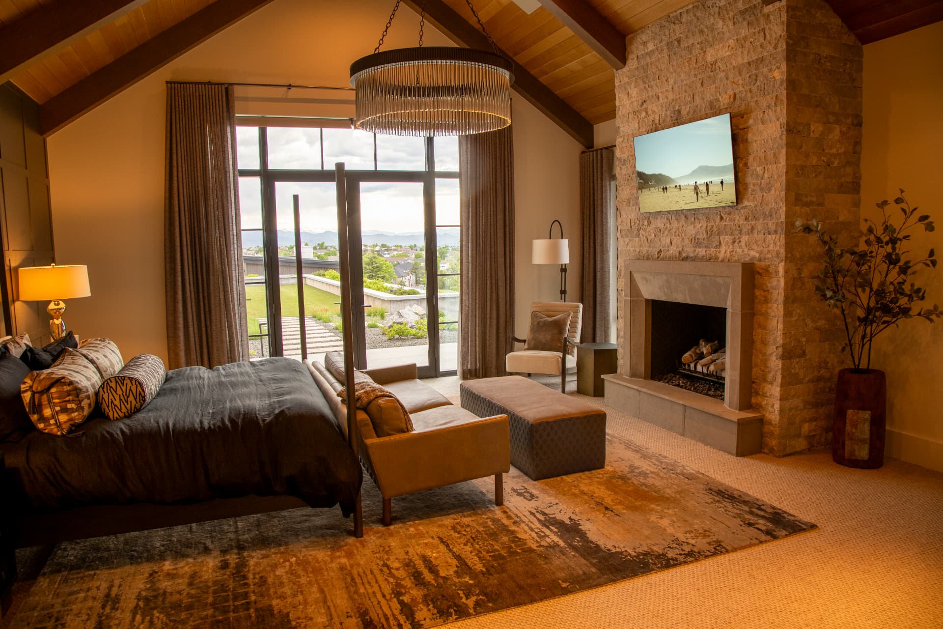 Primary suite with stone fireplace, wall-mounted television, and motorized drapery framing mountain views